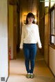 A woman in a white sweater and blue jeans standing in a hallway.