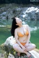 A woman in an orange bikini sitting on a rock by the water.